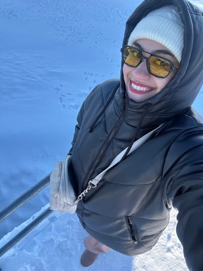A female subject is depicted from an upward-angled selfie perspective in a snow-covered environment. She wears a white ribbed knit beanie, black-framed sunglasses with yellow-tinted lenses, and a black hooded puffer jacket, zipped to the neck. A light beige crossbody bag with a silver chain link is slung across her torso. She is smiling, revealing white teeth and red lipstick. Her lower half shows light brown pants and brown, low-cut boots. The ground is uniformly covered in fresh snow, exhibiting numerous distinct human footprints. A metallic railing, possibly a handrail, is visible in the lower left quadrant, partially obscured by snow. The scene is illuminated by bright ambient light, indicating daytime conditions. The environment is consistent with the winter landscape of Uludağ mountain, located near Bursa, Turkey.