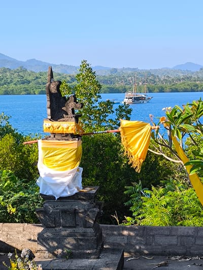 The image depicts a coastal or lakeside scene in Gerokgak, Indonesia, featuring a traditional stone shrine in the foreground, with a body of water, a boat, and forested hills in the background.
In the foreground, a dark grey, roughly textured stone shrine or altar stands centrally on a lower stone platform. The base of the shrine is wrapped in a plain white fabric, and above this, a golden fabric with a textured pattern and decorative border is draped. The upper part of the shrine is a dark, irregularly shaped stone carving with an upward-pointing element. A horizontal, reddish-brown wooden pole extends from behind the shrine towards the right. To the right of the shrine, a large, golden, fringed fabric object, resembling an umbrella or banner, is suspended from another wooden pole. Various green trees and shrubs occupy the mid-foreground, with some lighter green foliage appearing to the right of the shrine. The immediate foreground shows a dark grey stone wall or platform with scattered dried leaves and small patches of vegetation.
Beyond the foreground elements, a calm, deep blue body of water extends across the midground. A multi-masted wooden boat, consistent with a traditional Indonesian phinisi, is visible on the water to the left of the image's center. The boat has a white hull, dark accents, and visible rigging. The far bank of the water is lined with a dense expanse of various green trees and tropical foliage.
In the background, successive layers of green, tree-covered hills and mountains recede into the distance under a clear, pale blue sky. No human figures are visible in the scene.