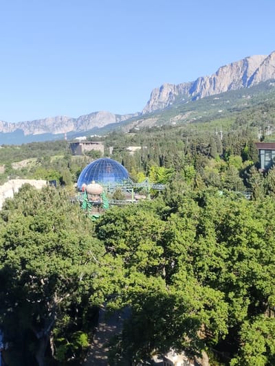 The image captures a daytime view in Симеизский поселковый совет, Ukraine, featuring a landscape dominated by lush greenery and mountains in the background. A prominent feature is a large, geodesic glass dome structure, possibly a building or attraction, surrounded by trees. In front of the dome appears to be a roller coaster or similar ride. The mountains in the distance are rugged and have varying shades of gray and brown. The sky above is clear blue. Scattered buildings are interspersed amongst the trees and hills. The foreground is largely filled with dense green foliage.