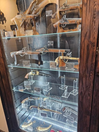 A display case with antique firearms is located in a store, likely a historical or souvenir shop, in Llandudno Junction, United Kingdom. The firearms include flintlock pistols, blunderbusses, and revolvers. Each firearm is displayed with a price tag and description, such as "18th Century Flintlock Pistol" or "1858 Percussion Black Revolver." The display case is made of wood and glass, and the firearms are arranged on acrylic stands. Some stands also display display stand, pistol keyrings, a Colt's bullet, and other items. The pricing shown indicates that these are probably reproductions.