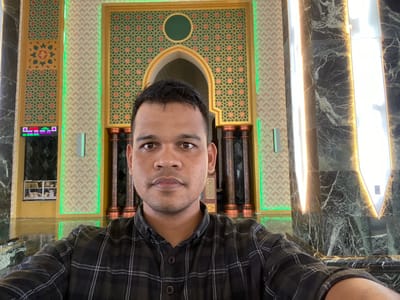 A man is taking a selfie in what appears to be the interior of a building with Islamic architectural elements. He is positioned centrally in the frame, facing the camera directly. He has dark hair, brown eyes, and is wearing a black and white checkered collared shirt.

The background features a large arched doorway adorned with intricate geometric patterns in green and orange, set against a beige textured wall. Tall, dark marble columns with prominent white vertical lighting flank the doorway. To the left of the doorway, a digital display showing times and possibly prayer schedules is visible, and further to the left, a glass-fronted cabinet containing books can be seen. The floor appears to be a highly reflective dark surface, possibly marble or polished stone, mirroring the surrounding architecture and lighting. The overall impression is one of a modern, well-lit interior space, possibly a mosque or a cultural center, located in the city of Aluekambuk, Indonesia.