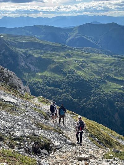 In Jošanica, Montenegro, three individuals are hiking on a rocky mountain trail. The path is strewn with light-colored stones and sparse green vegetation. The hikers, viewed from behind, are dressed in casual outdoor attire. The person in the foreground wears a plaid shirt, dark pants, a white cap, and a backpack. Two other hikers are further up the trail, one with a dark backpack and a white and black shirt, and the other with a blue backpack and a dark shirt, using a walking stick. The background displays a dramatic mountainous landscape with layers of green hills and peaks receding into the distance under a cloudy sky.