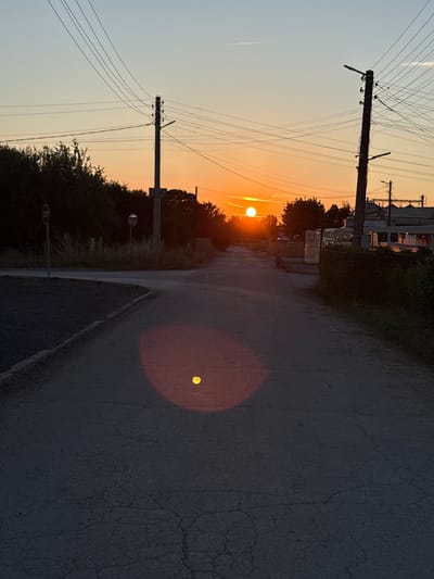 The image depicts a rural road scene in Petkovo, Bulgaria, at sunset. The sun is low on the horizon, casting a warm orange glow across the sky and illuminating the scene with golden light. The road, made of asphalt, is cracked and textured, with a faint white line marking its edge.
On the left side of the road, a power pole with numerous wires stretches towards the sky, silhouetted against the bright sky. Beyond the pole, trees and vegetation line the edge of the road, with some industrial structures visible in the distance. A sign with Cyrillic text, likely indicating a direction or place, is mounted on a pole.
The sun's reflection creates a bright, circular halo on the asphalt. The overall mood is serene and tranquil, with the warm sunset colors creating a peaceful atmosphere. The time of day is clearly sunset, indicated by the low position of the sun and the warm hues in the sky. There is no visible human activity in the image. The weather appears clear, with a faint contrail visible in the sky.