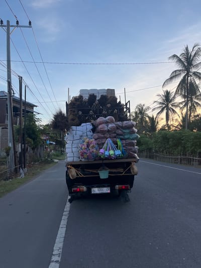 A black Isuzu truck is captured from behind, driving on a paved road in Blangtambue, Indonesia. The truck bed is heavily laden with various goods. Dominating the load are stacks of white plastic containers, bundled together, and numerous sacks of what appear to be produce, possibly coconuts, wrapped in plastic. Some of these sacks are reddish-brown, while others are green. Hanging from the back of the truck are several colorful plastic items, resembling small buckets or containers with handles. Bundles of dried grass or reeds are also visible at the top of the load.
The truck's license plate reads "BL 8244 ZR". In the background, to the left, a simple wooden house with a corrugated metal roof is visible, adjacent to a utility pole with multiple wires. To the right, lush green trees and palm trees line the road, with the sky displaying a gradient of blue and the warm hues of an approaching sunset or sunrise. The overall scene depicts a common mode of transportation for goods in a rural or semi-rural Indonesian setting.
