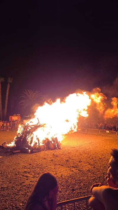 A large bonfire burns intensely in an open area at night, casting a bright orange and white glow on the surroundings. The fire is constructed from logs and other wooden debris. In the background, silhouetted palm trees are visible against the dark sky. A crowd of people stands behind a metal barrier, watching the bonfire. To the left, a red fire truck is parked. In the foreground, two individuals are partially visible, observing the spectacle. The ground is covered in gravel and sand. This scene likely depicts a traditional celebration or event, such as the San Juan festival, which commonly takes place in coastal areas like Almuñécar, Spain, where bonfires are a central element.