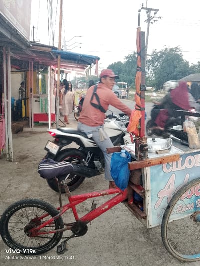 The image shows a street scene in Kecamatan Kroya, Indonesia. A man in a red shirt is riding a white motorcycle in the foreground. The man has a black strap across his chest and is wearing a black helmet. There is a red bicycle to the left of the motorcycle with a black seat and blue bag. The motorcycle is stopped near a food cart that has a blue and white sign and a large pot on top. There is a blurred motorcycle in the background with a person riding it. The man on the motorcycle is wearing a black hijab. There are people walking in the background and a building with a pink and white sign to the left of the image. The sky is cloudy and a utility pole with multiple wires and a large orange fabric is visible in the background.