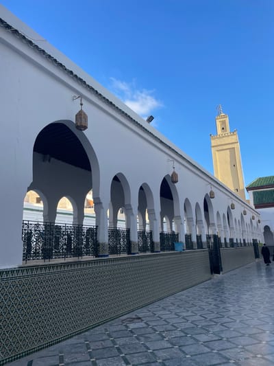 This image depicts the courtyard or exterior periphery of the Mausoleum of Moulay Idriss I in Moulay Idriss Zerhoun, Morocco, under a clear blue sky with scattered white clouds. A long, white-painted structure with a series of symmetrical horseshoe arches forms a colonnade along the left and central portions of the frame, receding into the background. The arches have dark undersides. Several ornate brass lanterns are suspended from the white wall between the arches. A decorative green scalloped trim runs along the top edge of the white structure. Below the arches, a black, intricately patterned metal railing with geometric designs is present. The lower section of the wall beneath the railing is adorned with elaborate green, white, and blue geometric mosaic tiles (zellige). The ground is paved with a grid of light and dark grey square and rectangular tiles arranged diagonally. In the background, a tall, light beige minaret with a square shaft and a decorative finial on its apex rises against the sky. To the right of the minaret, part of another building with a green-tiled roof is visible. One individual, dressed in a dark, flowing garment, is walking away from the viewer on the paved ground in the far right background. The scene is brightly illuminated by natural sunlight, casting minimal shadows.