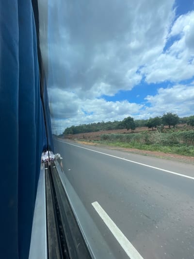 The image is taken from inside a moving vehicle, looking out the window. On the left, a dark blue curtain is visible, obscuring most of the interior. The window reflects a bright sky with scattered white clouds against a blue background.
The view outside shows a two-lane asphalt road stretching into the distance. The road has a white line marking the center. To the right of the road, the landscape is arid with sparse vegetation, including bushes and scattered trees with green foliage. The ground appears reddish-brown, suggesting dry soil. The overall environment appears rural and possibly a bit desolate. There are no people visible in the frame, nor any specific landmarks that would indicate the city of Sugali Tanda, India. The scene depicts a journey through a sparsely vegetated, rural area on a cloudy day.