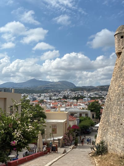 This is a vertical, eye-level, medium shot of a street in Rethymno, Greece, looking down a cobbled pathway lined with buildings and lush greenery. The sky is a vibrant blue with scattered white clouds.
In the foreground, a stone wall runs along the top of the frame. Below this, the scene opens up to a view of the town, with its white buildings and red-tiled roofs cascading down a hillside towards a mountain range in the distance. A pathway, paved with cobblestones, leads the eye into the scene. Several people are walking down the path. Some are seated at tables outside a cafe, which is adorned with potted plants and flowering bushes. A blackboard menu is propped up by the cafe. The time of day appears to be afternoon, given the bright sunlight casting shadows. The weather is sunny and clear, with a gentle breeze suggested by the presence of umbrellas. The overall mood is peaceful and inviting, suggesting a warm Mediterranean atmosphere. The architecture and landscape are characteristic of coastal towns in Greece. No visible text is present in the image.