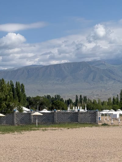 The image depicts a beach scene with a mountain range in the background, located in Koshkol', Kyrgyzstan. The foreground consists of a sandy beach. Behind the beach, a low, gray brick wall with visible mortar lines stretches horizontally across the frame. Several white beach umbrellas are visible, some appearing open and others closed, with their poles extending from the sand. A patch of green grass grows along the base of the wall. Beyond the wall, a line of tall, slender trees forms a dense green backdrop. Further behind the trees, a vast mountain range dominates the horizon, with its slopes appearing arid and rugged. The sky above is a light blue, dotted with scattered white clouds, some of which are fluffy and others more wispy. The overall scene suggests a peaceful, possibly resort-like, environment. There are no people visible in the image.