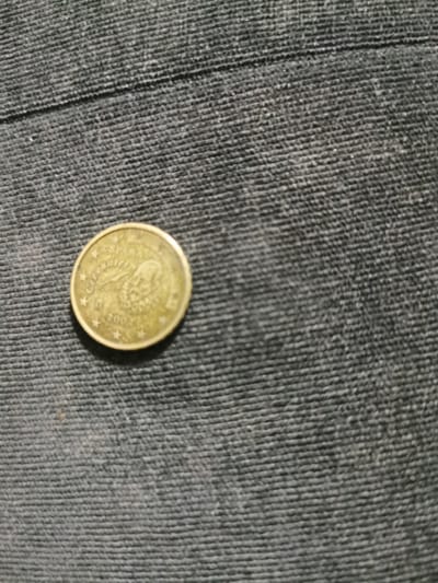 A single, gold-colored coin is lying on a dark, textured surface, which appears to be fabric with a visible ribbed pattern. The coin is circular and features an embossed design. The word "ESPAÑA" is legible at the top of the coin, indicating its origin is Spain. Below this, an image appears to be a portrait, possibly of a historical figure, with intricate detailing. The year "2001" is clearly visible towards the bottom of the coin, along with several small stars encircling the design. The coin is positioned slightly off-center to the left of the frame. The overall lighting is dim, creating soft shadows and highlighting the texture of the fabric. The coin is the sole subject in the image. The image was taken in Bourg-lès-Valence, France.