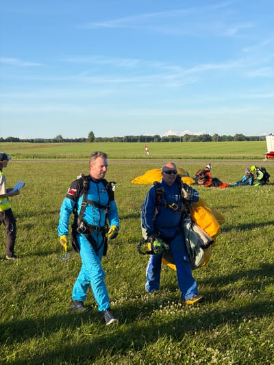 Two men in blue skydiving jumpsuits are walking across a grassy field under a clear blue sky. The man on the left is wearing yellow gloves and appears to be holding a clear piece of plastic. The man on the right is carrying a large yellow parachute. In the background, other individuals in skydiving gear are visible, along with scattered yellow parachutes and a white and red object. The scene suggests a skydiving event taking place in an open field, possibly near an airfield, consistent with locations like the airfield in Limbaži, Latvia, which is known for such activities. The overall environment is an outdoor recreational area with a focus on aerial sports.