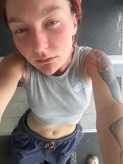 The media file shows a person in what appears to be a restroom or locker room. The person is wearing a gray tank top, dark blue pants, and a white athletic shoe. They have braided hair and several tattoos on their arm and shoulder. A small badge is attached to the tank top that reads "HOTEL KOH SAMUI." The background consists of dark gray walls and tiled flooring. The lighting suggests it could be daytime, though the interior setting makes it difficult to determine the exact time of day or weather conditions. The person is looking towards the camera, and their expression appears neutral. The image is taken from a close-up perspective, focusing on the person's face, upper body, and arm.