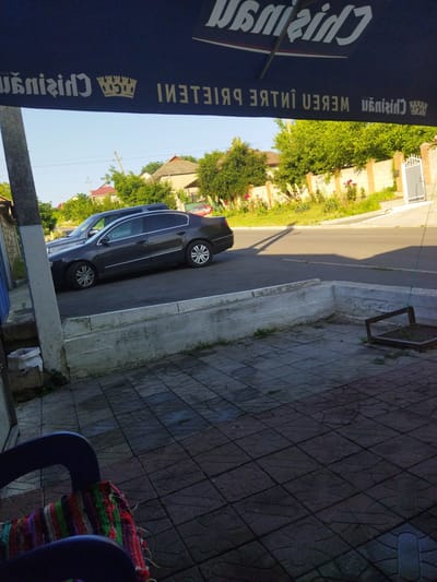 This is a medium-angle, outdoor, daytime photograph taken in Costești, Moldova. The scene is a street view, with a dark blue awning in the foreground casting a shadow over a paved area. The awning has white lettering and a crown logo. A dark-colored sedan is parked on the asphalt road in the middle ground, to the left of the frame. Behind the car and road, a fence made of brick and white metal posts runs horizontally. Beyond the fence, there are trees and houses. The ground in the immediate foreground is paved with square tiles. A blue chair with a colorful woven cushion is partially visible in the lower left corner.