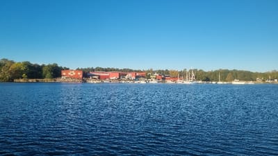 This panoramic image depicts a waterfront area in Västervik, Sweden. The lower half of the frame is occupied by a body of deep blue water with visible ripples, indicating a calm but dynamic surface. The middle ground showcases the developed shoreline. On the left, a dense line of green and partially autumnal trees forms a natural boundary, with a small, dark structure visible at the water's edge. Proceeding right, several large, red industrial-style buildings, consistent with warehouses or workshops, stand prominently on the shore. One such building displays a lighter square pattern on its facade. In front of these buildings, the shoreline appears rocky or reinforced, and a significant number of boats are present. These include numerous white recreational vessels, some with visible masts indicating sailboats, either docked in the water or stored on land along the embankment. The concentration of boats suggests a marina or shipyard area. Further to the right, the shoreline continues with more parked boats, transitioning back to a dense line of green trees. Faint outlines of smaller, lighter-colored structures are discernible within the trees on the far right. The upper half of the image is dominated by a clear, bright blue sky with no visible clouds. No human figures or discernible human interactions are present in the scene.