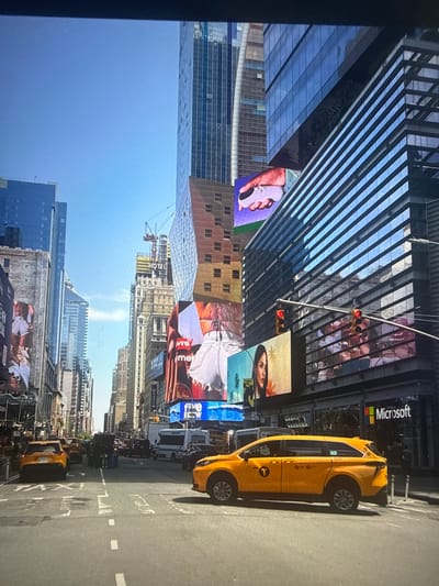 The image depicts a street scene in Times Square, New York City. Numerous tall buildings with digital billboards displaying various advertisements dominate the view. A yellow taxi cab is parked on the side of the street near a Microsoft store. Other vehicles and pedestrian traffic are visible further down the street. The sky is partly cloudy. There is no evidence that the image is taken in Des Plaines.