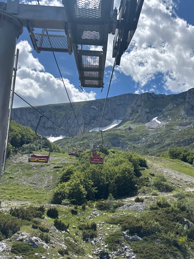 An upward-angle, medium shot captures a ski lift ascending a mountain in Motički Gaj, Montenegro. The ski lift's mechanical components, including pulleys and supporting structures, are prominent in the foreground against a sky filled with fluffy white clouds. Below, several ski lift chairs, some colored red and yellow, are suspended from cables, moving up the grassy, sparsely treed mountainside. The terrain is characterized by rocky outcrops, patches of green grass, and scattered shrubs. In the background, a large, imposing mountain face with visible snow patches rises, showcasing a rugged, natural landscape. The scene suggests a bright, clear day, likely during the summer or shoulder season, given the greenery and the presence of snow at higher elevations.