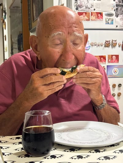 An elderly, bald male, wearing a pink polo shirt, is seated indoors, within a dining or kitchen area in Bnei Dror, Israel. He is actively consuming a small, round food item containing light-colored and green fillings, holding it with both hands and biting into it. His eyes are closed. A metallic watch is visible on his left wrist. On the table, positioned in front of the individual, are a clear glass containing a dark, chilled beverage with condensation, and a mostly empty white plate with an embossed pattern. The table surface is covered by a light-colored tablecloth adorned with a repeating black eye motif. In the background, a white refrigerator door is visible, decorated with numerous magnets, including a series of owl figures, various personal photographs, and a colorful heart-shaped design.