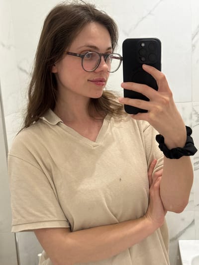 The scene depicts a young adult female capturing a mirror selfie in an indoor setting within Scafati, Italy. The woman, shown from the mid-torso up, has long brown hair and wears round, dark tortoiseshell-framed eyeglasses. She is dressed in a light beige V-neck short-sleeved top, which exhibits a small, dark mark on the front, slightly below the center of her chest. Her left wrist is adorned with a black scrunchie, and her left hand holds a black smartphone, featuring a triple-lens camera system and a visible circular MagSafe attachment on its case, positioned to capture her reflection. Her right arm is bent, with the forearm resting across her abdomen. The background consists of a white tiled wall with subtle gray veining, characteristic of a bathroom setting, with a vertical tile seam visible on the left and a partial view of a white surface, possibly a sink or counter, visible in the bottom right.