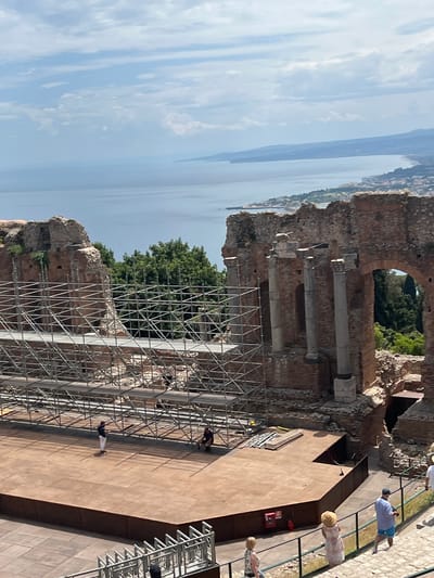 The image shows a view from the upper seating area of the ancient Greek Theatre of Taormina, Italy. The theatre's stage area, which is currently set up with wooden platforms and scaffolding, is visible in the foreground. Several people are present on and around the stage area, some appearing to be workers based on their attire and activities. The iconic ruins of the theatre's structure, with brick walls and columns, are prominent. Beyond the theatre, a panoramic view of the sea, the coastline of Taormina, and the distant mountains under a cloudy sky is displayed. The scene suggests ongoing work or preparation for an event at the historical site.