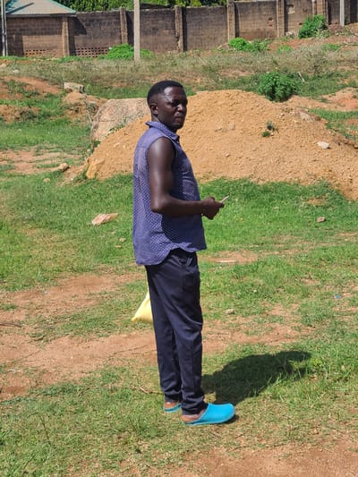 A dark-skinned man is standing in a grassy, dirt-filled outdoor area, possibly in the city of Barikin Jirga Summa, Nigeria. He is looking to his right and holding a silver object, likely a phone, in his right hand. He is wearing a dark blue sleeveless shirt with white polka dots and dark navy pants. His feet are clad in bright blue slip-on shoes with brown soles. In front of him, a bright yellow bag is partially visible on the ground. The background features a dirt pile, rough grass, and a brick wall with a damaged section.