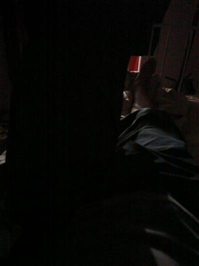 The image is very dark, with limited visibility. It appears to show a person lying down, covered by dark fabric, possibly a blanket or sheets. The lighting is dim, suggesting it might be nighttime or a dimly lit room. There are hints of other objects in the background, including what looks like a red curtain or fabric, and a vertical structure that could be part of a window or door frame. The overall impression is of a private, indoor setting. The person is not clearly visible, making it difficult to determine their age, gender, or any specific features. The image lacks any clear indication of the activity taking place, beyond a person resting or sleeping. There is no visible text in the image. The location context of Juma, Uzbekistan, does not provide any discernible cues within the visual information of the image itself.