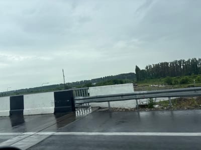The image captures a view from a vehicle on a wet, grey road, likely after rain, in the city of Lanchkhuti, Georgia. The road surface is reflective, showing lane markings. A concrete barrier, painted with alternating black and white stripes, runs along the side of the road, separating it from a metallic guardrail. Beyond the guardrail, there is a grassy embankment with some scattered debris and a dense line of trees forming a backdrop. In the distance, a cell tower is visible against a cloudy, overcast sky. The overall scene suggests a rural or semi-rural setting within or near Lanchkhuti.
