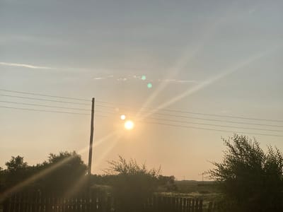This media file captures a rural scene in Rakity, Russia, during what appears to be sunset or sunrise. The sky is hazy and a pale yellow-orange, indicating either dusk or dawn. Three bright orbs are visible in the sky: the main sun, which is a bright, diffused circle with strong light rays emanating from it, and two smaller, greenish orbs, possibly lens flares or atmospheric phenomena.
The scene is dominated by a power pole with multiple wires stretching across the frame, partially obscuring the sky. In the foreground, a dark wooden fence runs horizontally. To the left and right of the frame, silhouetted trees and bushes add depth to the landscape. The trees are dense and leafy, with sunlight filtering through their branches, creating bright highlights. In the distance, a flat, open field is visible, with a few more power poles dotted across it, suggesting a rural or semi-rural setting.
The overall mood is serene and peaceful, with the soft, diffused light creating a tranquil atmosphere. There is no visible text in the media file. The presence of power lines and the open landscape are characteristic of a Russian countryside. The time of day is suggested by the low angle of the sun and the warm, hazy light. The weather appears calm, with no strong winds evident.