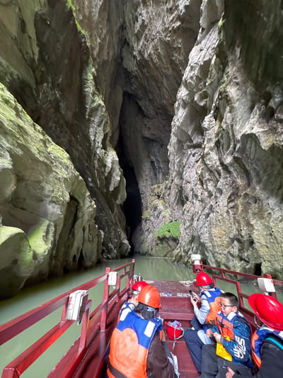 The image shows a boat tour inside a cave near Zhuoshui, China. On the right side of the image, people wearing life vests and red helmets are on a boat. The boat has a red metal railing and what appears to be white plastic boxes affixed to the railing. The left side of the image shows the cave wall. The cave walls are jagged and rocky, with some green moss. In the center of the image, there is a dark opening further into the cave.
