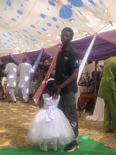 An outdoor celebratory event in Kerang, Nigeria, is depicted under a large, temporary canopy. The canopy, primarily white, features numerous circular cut-outs through which a clear blue sky is visible, indicating bright daylight and fair weather. Decorative fabric drapes in shades of purple, white, and pink adorn the canopy's structure. The ground is covered with dried straw or grass, with a section of green mat visible on the right.
In the foreground, a tall man wearing glasses, a dark short-sleeved collared shirt (with faintly visible white text, possibly "BIFT," on his right chest pocket), and dark cargo pants, looks towards the camera. He has short, dark hair and gently places his right hand on the head of a young girl beside him. The girl is adorned in a beautiful white party dress featuring a full, ruffled tulle skirt, a floral-patterned bodice, and a large white bow tied at her waist. She wears a white headband over her dark, curly hair, and her face is turned away from the camera, looking downwards or to the side.
In the background, several other attendees are visible. A person in a green garment is partially seen behind the man. Further into the scene, two men dressed in white, long-sleeved traditional robes or suits are standing, with additional people seated further back. Floral arrangements are visible on a table, contributing to the festive and communal atmosphere of the gathering, which appears to be a formal event such as a wedding or dedication ceremony.