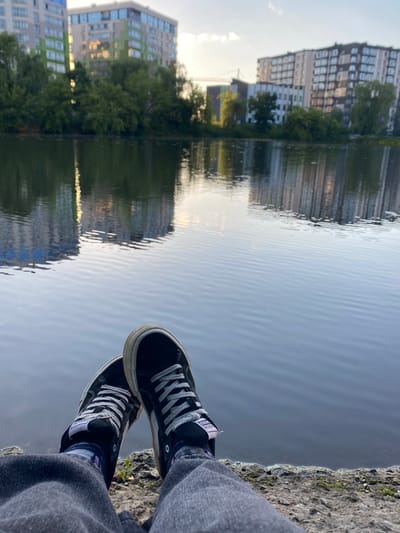 The image shows a person's legs and feet, wearing black sneakers with white laces and grey jeans, resting on a concrete edge next to a body of water. The water reflects buildings and trees, indicating an urban environment. The architecture visible in the reflection suggests residential buildings, likely apartment complexes. The presence of water and urban structures implies a location such as a waterfront park or a residential area adjacent to a lake or river. Given the context of Ivano-Frankivsk, Ukraine, this scene could be depicting a view from the embankment of the Bystrytsia-Solotvynska river or a similar urban water body within the city. The time of day appears to be late afternoon or early evening, with soft lighting and calm water. There are no other people or animals clearly visible in the frame.