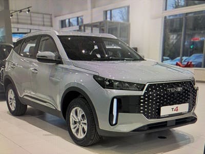 A silver TENNEL T4 SUV is parked indoors, likely in a car dealership showroom, in Mirnoye, Ukraine. The vehicle has a large, textured grille with a TENNEL emblem above a T4 badge. Vertical LED lights are integrated into the front bumper. The car is viewed from the front-left angle, with its tires and side profile visible. The showroom floor is tiled in a light color. In the background, through large windows, other cars can be seen, including what appears to be a red vehicle and a blue one. The lighting suggests an indoor environment, possibly with natural light filtering through the windows. There are no people visible in the image.