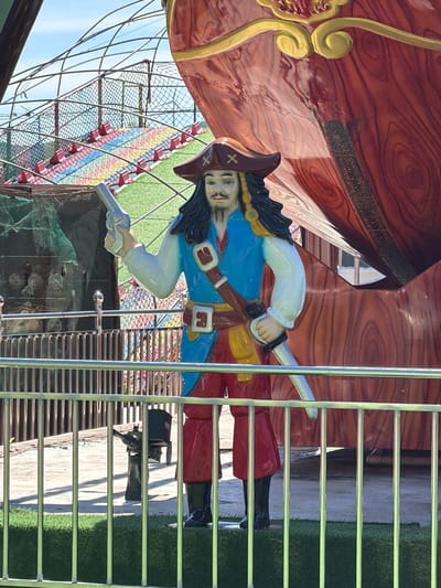 The image displays a colorful pirate figure, likely a statue or prop, positioned within what appears to be an amusement park or play area. The pirate, centrally located, is depicted in a dynamic pose, holding a silver sword in one hand and a silver pistol in the other. He wears a blue jacket, a white ruffled shirt, brown belts with gold buckles, white breeches, black boots, and a dark brown tricorn hat marked with a white 'X' symbol. His facial features include a dark goatee and a slight smile.

The pirate is set against a large, reddish-brown structure designed to resemble a wooden ship's hull, complete with painted wood grain and ornate gold scrollwork details. This structure has a glossy, reflective surface. To the right, a metal railing or ladder with numerous horizontal bars extends from top to bottom, partially obstructing the view of the pirate and the background.

Beyond the railing, artificial green turf is visible on the ground, suggesting an outdoor setting. In the background, under a bright blue sky, there is a large, multi-colored structure that looks like a climbing net or play apparatus, featuring blocks or ropes in red, yellow, blue, and green hues, enclosed by dark netting. The scene is brightly lit, indicating a clear, sunny day. The overall impression is of a vibrant, themed environment designed for entertainment.