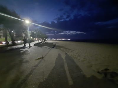 This image depicts a nighttime scene along the coast in Atakum, Turkey. A bright street lamp illuminates a paved walkway bordering a sandy beach and the sea. Two figures, likely human, are casting long shadows on the walkway. In the background, cars are visible, suggesting the presence of a road or parking area adjacent to the walkway. The sky is dark blue with some scattered clouds, and a distant, blurred line of lights suggests a more populated area or cityscape along the coast. A small, indistinct object lies on the walkway in the middle ground, possibly a piece of debris. The overall atmosphere is tranquil, with the artificial light from the lamp creating a dramatic contrast with the dark sky and sea.