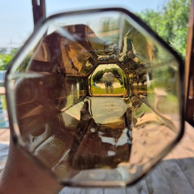 The image is a close-up, distorted view looking through a clear glass, likely a drinking glass, from the inside. The glass has an octagonal shape. The background seen through the glass is an outdoor environment, appearing to be a garden or yard. Sunlight filters through, creating reflections and highlights on the glass. The distant background shows green foliage and a bright sky. The glass is held by a person's hand, which is visible on the left side of the frame, partially obscuring the view. The overall impression is that of a casual, possibly domestic setting, taken from a low angle. The location in Новостройка, Russia is not discernible from the image itself; it is an inferred detail.