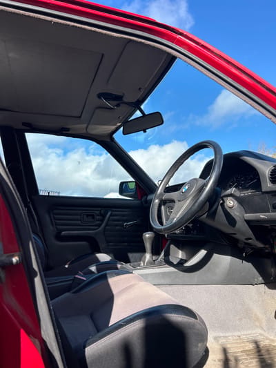 The image shows the interior of a red car, possibly parked. The door is open, revealing a gray interior with a black dashboard, steering wheel with a BMW logo, and gear stick. A rearview mirror is visible, and the blue sky with clouds is reflected in the windshield. The location of the image is inside the vehicle and the city is not visible. There are no people in the shot.