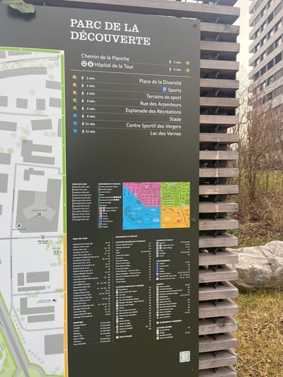 The image displays an outdoor scene, slightly tilted, focusing on a large, dark gray informational sign. The sign is titled "PARC DE LA DÉCOUVERTE" (Discovery Park), serving as a guide for visitors in Meyrin, Switzerland. Behind the sign, a wooden fence runs horizontally, and in the background, there are trees with bare branches, suggesting late winter or early spring, and modern multi-story buildings, indicating an urban or suburban setting. The scene is illuminated by daylight, implying clear or bright overcast weather.

No people are visible in the frame; the primary subjects are the sign, the fence, and the distant architecture and greenery. The sign itself is the central object of interest, providing detailed information about the park and its surroundings.

The left section of the sign lists various destinations with estimated walking times and directional arrows. These include "Chemin de la Planche" (1 min), "Hôpital de la Tour" (1 min, with a hospital icon), "Place de la Diversité" (1 min), "P Sports" (Parking Sports), "Terrains de sport" (3 min), "Rue des Arpenteurs" (5 min), "Esplanade des Récréations" (6 min), "Stade" (11 min), "Centre Sportif des Vergers" (11 min), and "Lac des Vernes" (11 min).

The right side of the sign features a colored map divided into four main zones (green, yellow, pink, and blue), accompanied by a comprehensive legend and directory. Categories listed include "ÉQUIPEMENTS ET SERVICES" (Equipment and Services), "ACTIVITÉS CULTURELLES ET SOCIALES" (Cultural and Social Activities), "ACTIVITÉS SPORTIVES ET LUDIQUES" (Sports and Leisure Activities), "TRANSPORTS PUBLICS" (Public Transport), "ACTIVITÉS ET COMMERCES" (Activities and Shops), and "PISTE CYCLABLE" (Cycling Path). An "INDEX DES VOIES" (Index of Roads) provides a list of street names with corresponding map coordinates, alongside a section for "QUARTIERS" (Neighborhoods). A QR code is present in the upper right corner of the sign.