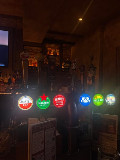 The image shows a dimly lit bar with several beer taps and illuminated logos. In the foreground, a menu titled "COLD BEER cutting my name" is partially visible, displaying categories like "FROM THE TAPS," "FROM THE FRIDGE," and "FROSTY." The beer taps feature logos for Amstel Bier, Heineken Original, Hickory's Smokehouse Pale Ale, Bud Light, Sierra Nevada Pale Ale, and Driftway Tropical IPA. In the background, bottles of alcohol are visible on shelves, and the overall ambiance suggests a rustic or traditional pub setting. There are no people visible in the image. The image does not provide any information to determine its location in the city of Peterborough, United Kingdom.