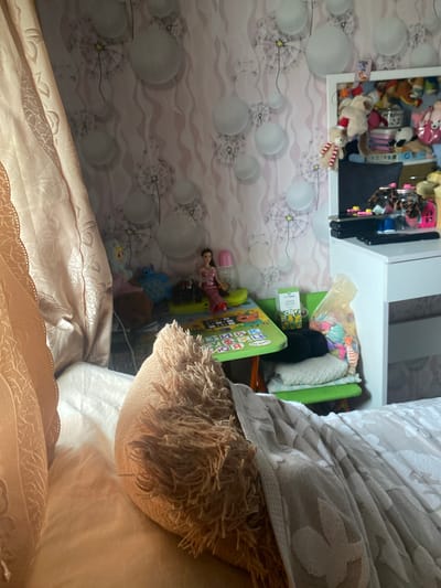 The image depicts a bedroom interior. In the foreground, a fluffy, beige pillow rests on a white bedspread with a floral pattern. To the left, a tan curtain with a floral pattern hangs down. The background features light pink wallpaper with a dandelion motif. A small green table with various toys and a doll is visible. A white dresser with shelves full of stuffed animals and other items stands to the right. A plastic bag filled with colorful toys is on a green chair next to the table. The location is a bedroom in the city of Kamenka, Russia.