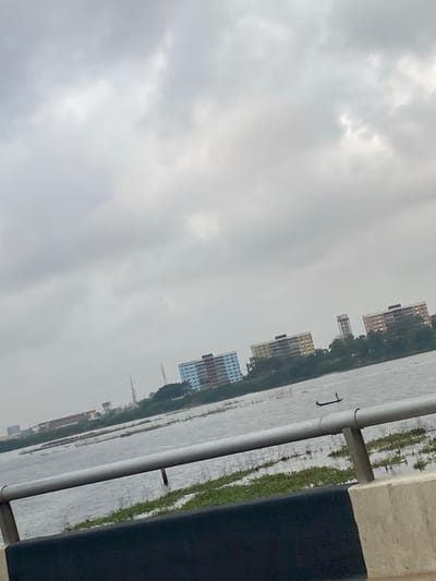 The image captures a panoramic view across a body of water in Ebute-Metta, Nigeria, observed from what appears to be a bridge. The sky is uniformly overcast, filled with light grey clouds. In the immediate foreground, a dark grey concrete barrier topped by two light grey metallic horizontal railings extends across the lower portion of the frame. The water below the railings shows moderate surface ripples and contains numerous scattered patches of green aquatic vegetation. In the mid-ground, a single dark-colored, narrow boat or canoe navigates the water, occupied by one individual standing upright, facing towards the left. The distant shoreline, beyond the water, is lined with various structures. Prominently, three multi-story buildings are visible: one with light blue and brown sections, a central one with yellow and brown sections, and a rightmost one with light brown and orange sections. A tall, slender water tower stands behind the rightmost buildings. Further to the left along the distant shore, multiple industrial structures, including transmission towers, are discernable amidst sparse trees and distant urban development.