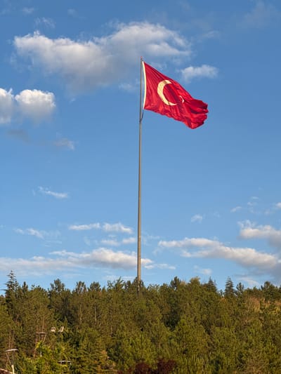 A tall flagpole stands prominently in the foreground, a Turkish flag with a white crescent and star on a red background is flying from it. The flag is positioned in the upper right portion of the image and is illuminated by sunlight. The flagpole extends vertically through the center of the frame. Below the flagpole and the flag, a dense line of green trees forms the middle ground, with some of the trees appearing taller and more spread out than others. In the lower part of the image, the tops of streetlights and some undergrowth are visible, suggesting an urban or semi-urban park setting. The background is a clear blue sky with scattered white clouds. The image appears to have been taken in Gölbaşı, Turkey, indicated by the context.