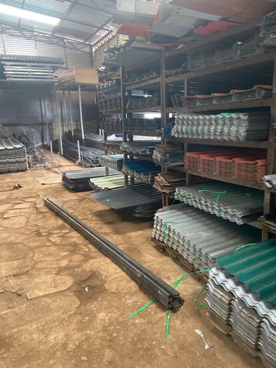 The image depicts the interior of a building in Biashara Ward, Kenya. The space appears to be a storage area or shop selling corrugated metal sheets and metal beams, possibly for construction. Stacks of various colored corrugated sheets (green, silver, red, blue) are stored on shelves constructed of metal frames. The floor is a rough, textured surface, possibly concrete or packed earth. A bundle of metal beams tied with green twine is visible on the floor in the foreground. The area is lit by natural light filtering in from an unseen source, possibly a doorway or window to the left. No people are visible.
