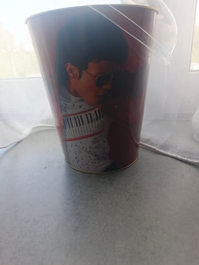 A cylindrical metal container, possibly a popcorn bucket, is depicted in the image. The container features a prominent graphic of a person, likely Michael Jackson, wearing sunglasses and a red jacket. Below the image of the person, there is a design of a piano keyboard and musical notes. The container has a clear plastic lid and a clear plastic handle attached to the sides.
The container is placed on a flat, grey surface, which appears to be a table or countertop. In the background, a white, sheer curtain is visible, suggesting the container is near a window. The lighting suggests it is daytime, possibly indoors, with natural light filtering through the curtain. The overall scene is still and inanimate, with no discernible activity taking place beyond the static display of the object. No specific time of day, weather conditions, or strong emotions are evident. No visible text is present on the container or in the immediate surroundings. The location context of Nakhabino, Russia, is provided but not visually indicated within the image itself.
