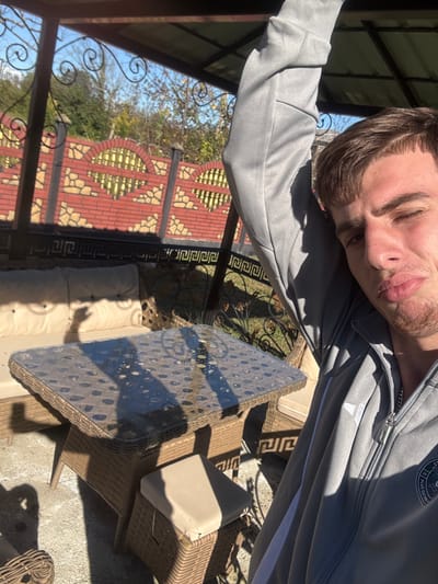 The image depicts a male individual taking a selfie in an outdoor setting within Durgena, Georgia. The subject, positioned in the foreground and right side of the frame, is a male with short brown hair and light stubble, estimated to be in his mid-20s. He wears a grey zip-up jacket with white stripes on the sleeves and a dark emblem on the left chest, and a silver chain is visible around his neck. His right arm is raised, and his left eye is closed in a wink, with lips slightly pursed towards the camera.

The scene is set under a dark metal gazebo with a corrugated roof, partially visible at the top and left. In the midground, a rectangular dark brown rattan-style outdoor table with a clear glass top is present, exhibiting sunlight reflections and shadows. Behind the table, a light-colored cushioned rattan bench is visible, and a smaller square cushioned stool is partially seen under the table. The ground consists of a light-colored paved surface.

In the background, a decorative fence is prominently featured. It is constructed from red brick pillars and sections, incorporating a stone base and arched openings with golden/yellow horizontal slats behind them, alongside diamond-patterned brickwork. Dark metal decorative elements with a Greek key pattern are at the fence's base. Beyond the fence, green trees and bushes are visible under a clear blue sky, indicating a sunny day.