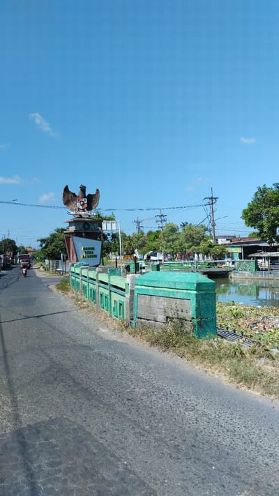 The image captures a street scene in Plumbungan, Indonesia, dominated by a large monument featuring the Garuda Pancasila, the national emblem of Indonesia. The monument is positioned on the left side of the road, with text below it that appears to read "PASUK DESA BAMB..." which likely signifies a village name. To the right of the monument, a canal or river runs parallel to the road, bordered by a green, ornate fence. Lush green trees are visible in the background, interspersed with utility poles and wires. On the road, a motorcycle is in motion, indicating movement. The sky is a clear, bright blue with a few wispy clouds, suggesting a sunny day. The road surface is asphalt, showing some wear and tear. The overall environment appears to be a semi-urban or rural area within Plumbungan.