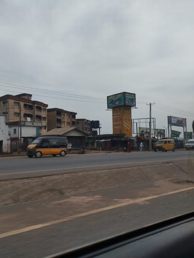 The image presents an urban scene in Ogbunike, Nigeria. A road runs horizontally through the foreground, with a gray and cloudy sky overhead. On the opposite side of the road are several buildings, including multi-story residential structures and smaller shops. Traffic consists of commercial vehicles, including vans painted in yellow and black. A billboard displaying advertisements for 'West Lake' tires and 'Maxxis' tires is visible, indicating a commercial district. There are also some individuals present near the shops, but the image does not reveal any specific actions or interactions.