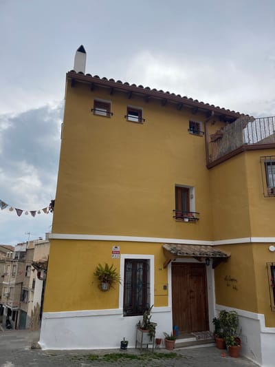 The image depicts the exterior of a three-story building in Buñol, Spain, under an overcast sky. The building features a prominent mustard-yellow facade with a white painted base. Its roof is tiled with reddish-brown semicircular barrel tiles, from which a white, rectangular chimney with a pointed top ascends on the left side. Dark brown wooden beams support the roof overhang.
The building's left section presents a series of vertically aligned windows: two small, rectangular windows on the top floor, a taller rectangular window on the middle floor, and a large rectangular window on the ground floor. All visible windows feature dark brown frames and external dark brown metal railings; the middle-floor window holds two small potted plants, and the ground-floor window is covered by vertical metal grilles and has a decorative wrought iron cage containing a green fern attached below.
The main entrance, located towards the center, consists of a double-leaf, dark brown wooden door with a small, gabled canopy of terracotta tiles overhead. A small, patterned mat lies at the doorstep. The house number "14" is affixed to the wall to the right of the ground-floor window, beneath a yellow utility box. A red and white "Alarma PTS" sign is mounted on the wall to the left of the ground-floor window. Numerous potted plants, including terra cotta and grey planters, are arranged around the main door and ground-floor window.
To the right of the main facade, a recessed section of the building includes a balcony with a metal railing and a bamboo screen, and a window below it. The ground in front of the building is paved with light grey, irregular flagstones. To the left, a narrow, uphill street is visible, flanked by other residential buildings of various heights. A string of colorful, patterned banners hangs across the street in the upper left background. No people are visible, and no actions or interactions are depicted.