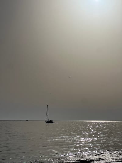 The image captures a coastal scene near Almancil, Portugal, characterized by bright, hazy atmospheric conditions. The sun, a prominent luminous disc, is positioned in the upper right quadrant of the frame, casting intense white glare across the water surface, particularly on the right side. In the mid-ground, a dark-silhouetted sailboat with a single mast is stationary, anchored or moored slightly to the left of the image center. Further in the distance, towards the left, two smaller, dark-silhouetted vessels are faintly visible on the water. A small, dark-silhouetted airplane is depicted in flight in the upper mid-right portion of the uniformly light grey-brown sky. The water in the foreground exhibits subtle ripples, and dark, undefined masses are present along the very bottom right edge, possibly indicating rocks or shoreline. No individuals are discernible within the frame.