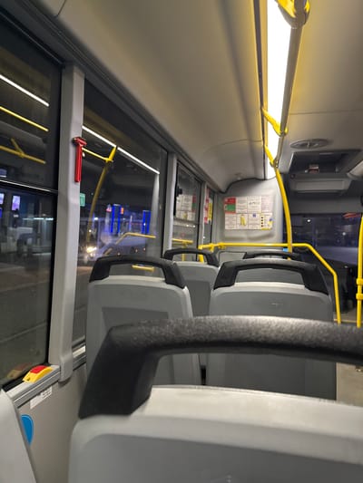 The image depicts the interior of a bus, likely in motion, featuring a row of gray seats with black handle supports. Yellow handrails run along the interior structure. A red emergency hammer is visible near the window. Information signs are affixed to the wall near the front of the bus. The window view suggests an urban setting at night, potentially in Trekhgorka, Russia. Lights from buildings and other vehicles are visible through the window. The scene is devoid of people except that there are the heads of two blurred people through the window.