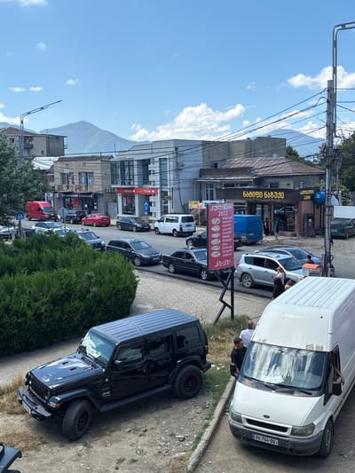 In Khashuri, Georgia, on a sunny day with a partly cloudy sky, a street scene unfolds with a focus on vehicles and buildings. A black Jeep Wrangler Sahara is parked on the left, its hood raised slightly. In front of it, a white Ford Transit van is parked, its windshield streaked with dirt and reflections. Two men are visible near the van; one, wearing a dark shirt, stands by the open driver's side door, while another man in a white shirt stands behind him.
The street is lined with various cars, including a red sedan, a dark SUV, and a dark sedan. A tall green hedge occupies the left foreground. Buildings flank the street, featuring a mix of architectural styles. One building displays a large red sign with Georgian text, and another, with a brown awning and yellow lettering, appears to be a food establishment. Power lines crisscross the sky above the scene. In the background, a verdant mountain range is visible under the bright sky.
